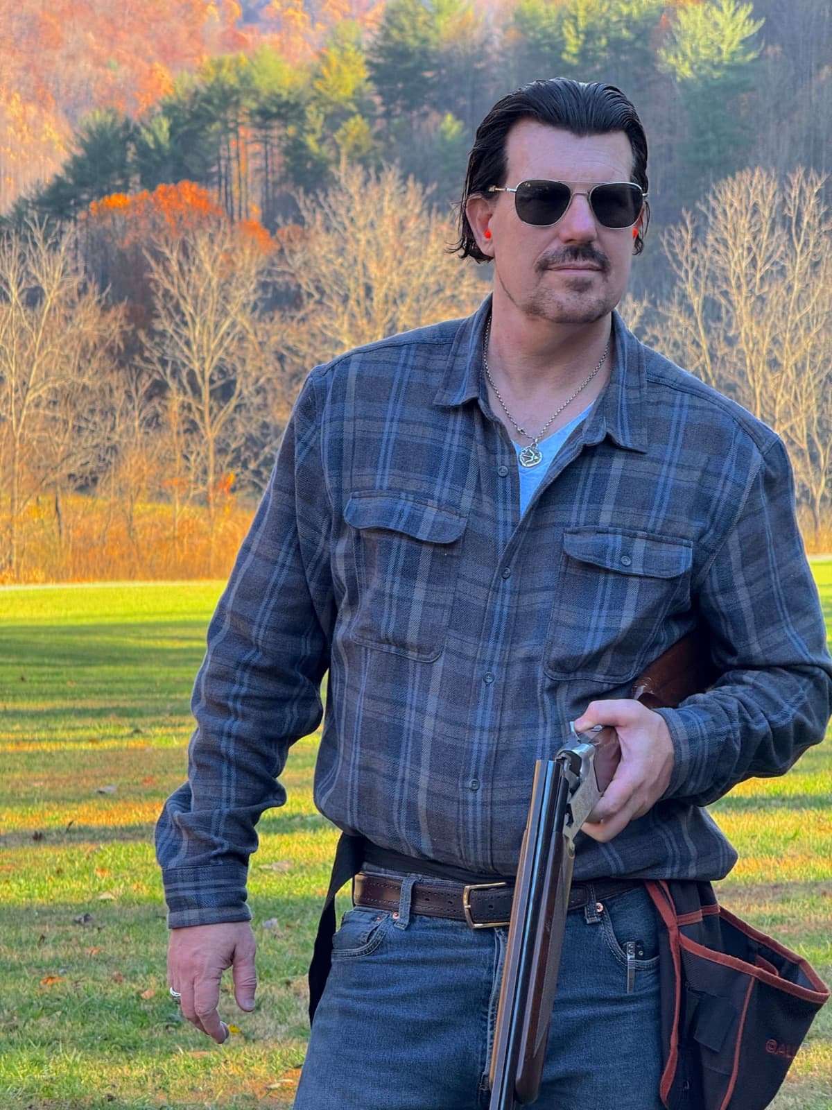 Chuck Pierce outdoors holding a shotgun, with autumn trees behind him.